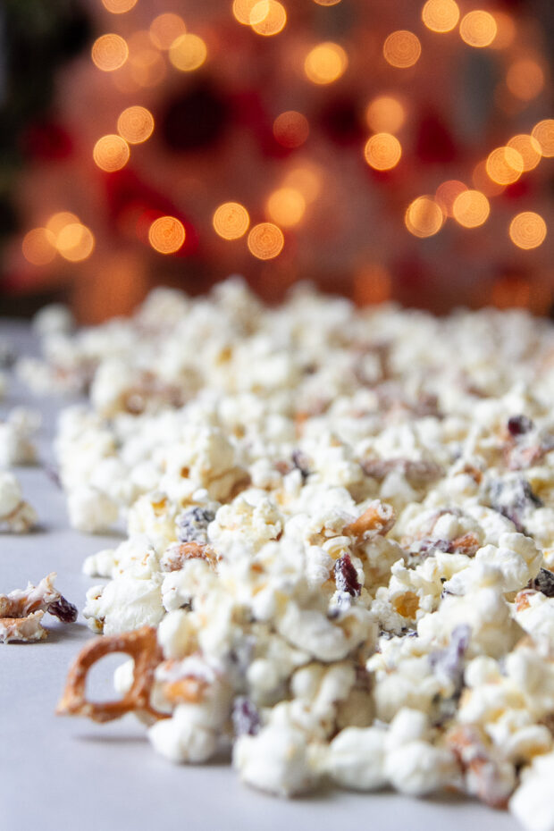 White Chocolate Popcorn with cranberries, pretzels, & orange laying out on parchment to set with a Christmas tree in the background.