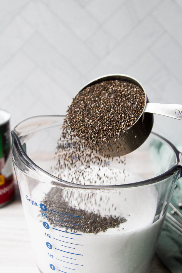 A measuring cup pouring chia seeds into a large, clear liquid measuring cup filled with coconut milk and water.