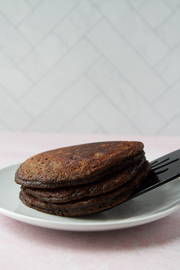 A stack of three chocolate paleo pancakes placed on a plate with a spatula.