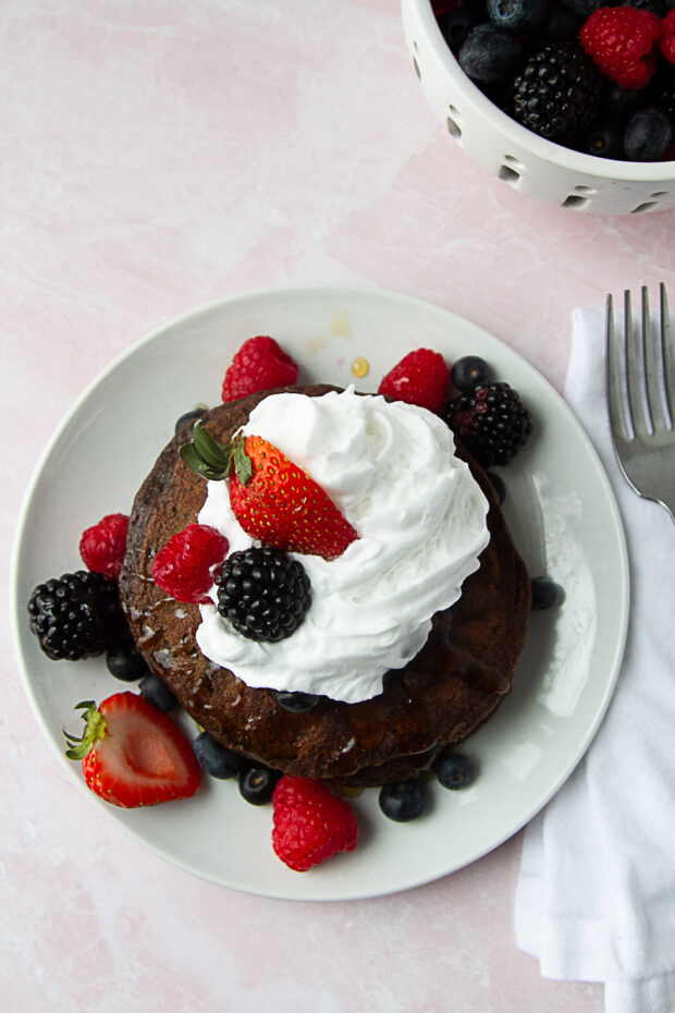 Dark Chocolate Paleo Pancakes on a white plate topped with coconut whipped cream, fresh berries, and a drizzle of honey.