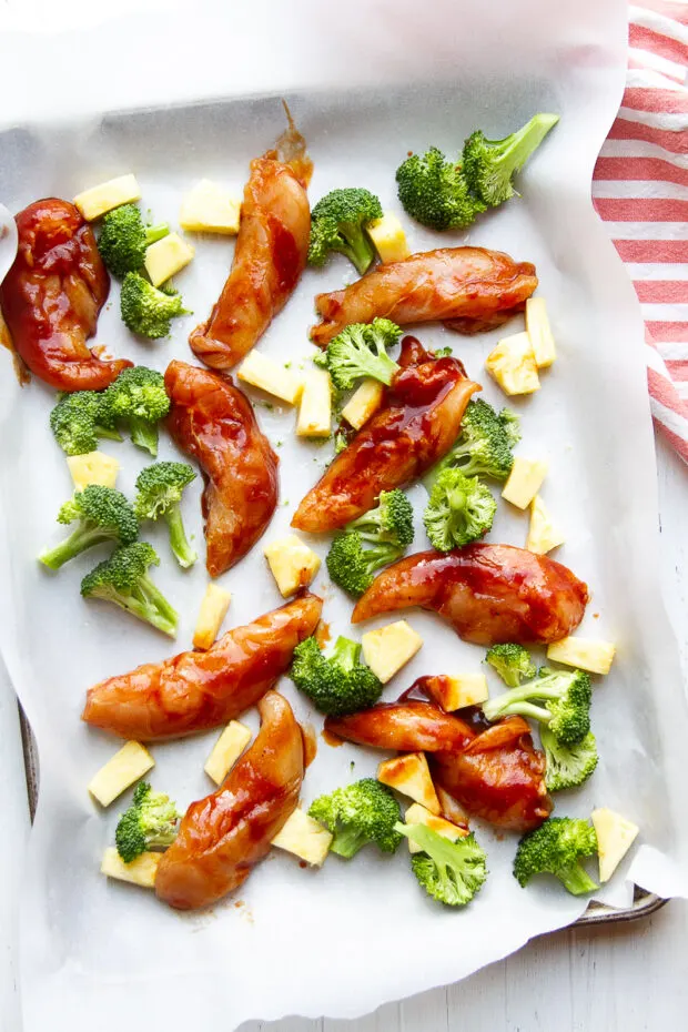 Parchment lined baking sheet with sauce-coated chicken tenders, raw broccoli florets, and fresh pineapple chunks.