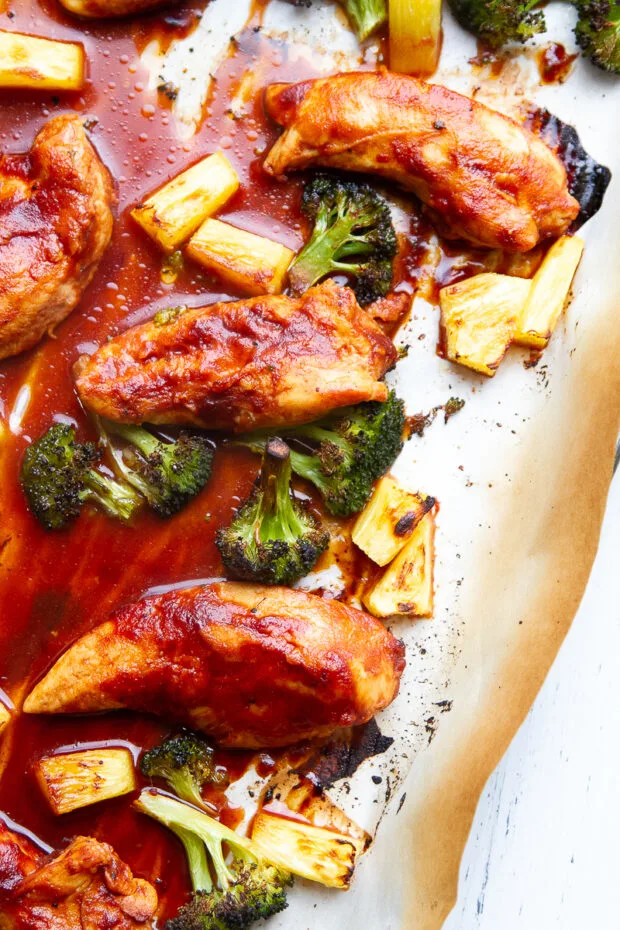 Cooked chicken tenders broiled and brushed with BBQ sauce on a parchment lined sheet pan with caramelized pineapple & broccoli.