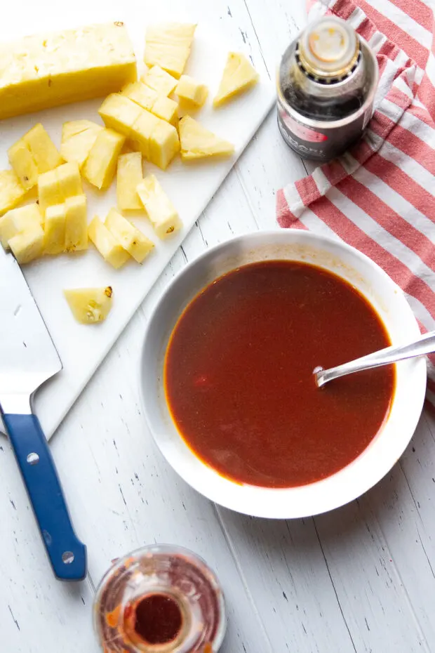 BBQ sauce mixture stirred together in a white bowl. Cutting board with chopped fresh pineapple.