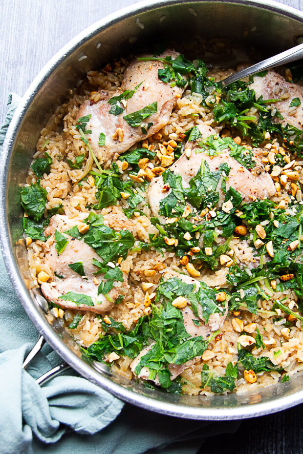 Skillet with finished chicken thighs, coconut rice, topped with cooked kale and crushed peanuts.