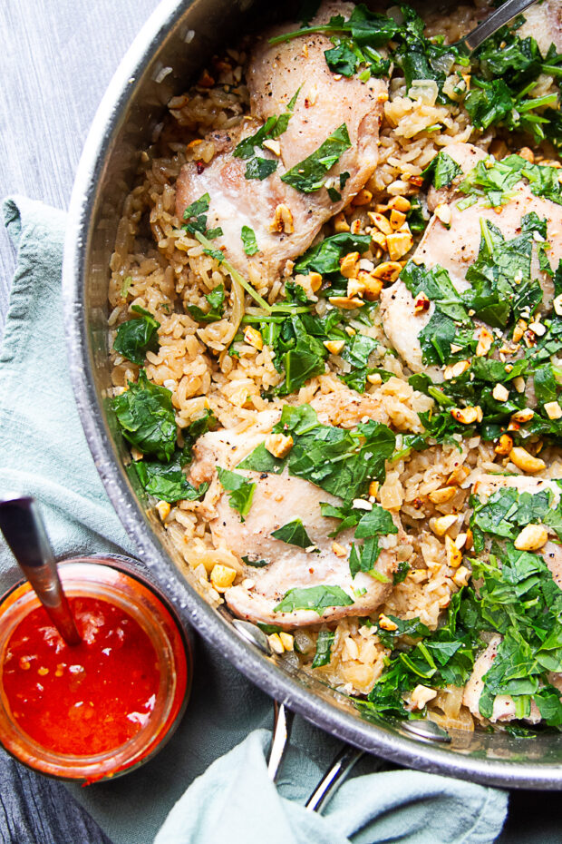 Skillet with finished chicken thighs, coconut rice, topped with cooked kale and crushed peanuts. Asian chili sauce on the side.