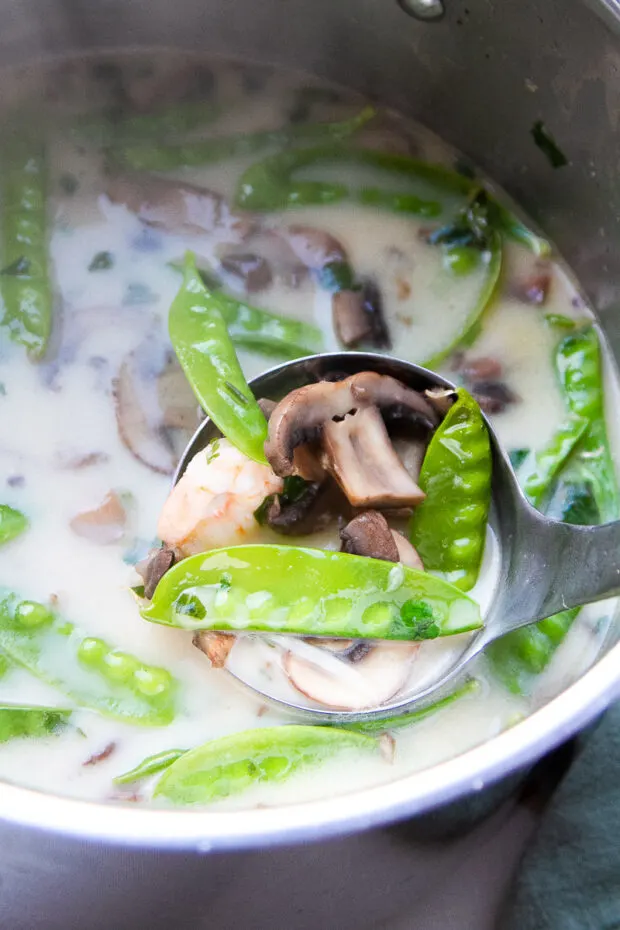 View of inside the pot -- a ladle of soup being lifted out with shrimp, sliced mushrooms, snow peas.