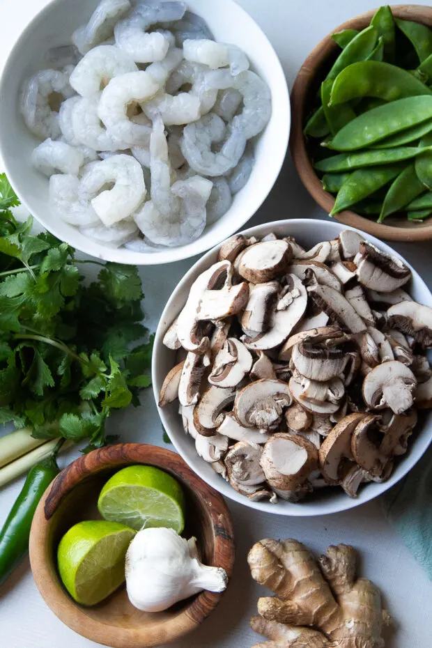 Bowls of ingredients -- sliced mushrooms, uncooked shrimp snow peas, limes, garlic, ginger, fresh cilantro, lemongrass, Serrano chili.