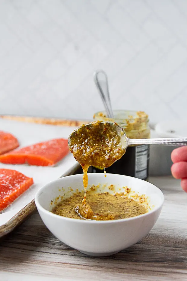 Same bowl with the honey mustard sauce with the spoon lifted as the sauce slides off. Salmon fillets visible in the background.