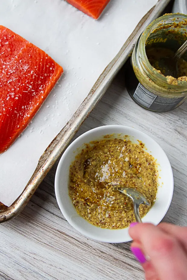 A small bowl with whole grain mustard, honey, and garlic being stirred together. Salted raw salmon on a sheet pan is off to the side.