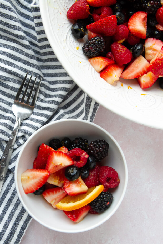 A serving of berry salad in a small white bowl next to the serving bowl with the rest of the salad.