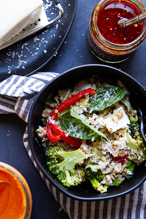 Black bowl with a serving of the Italian Chicken Sausage Skillet. Shows block of fresh parmesan and chili crunch off to the side.