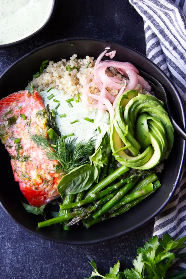 Black bowl with roasted salmon, asparagus, quinoa, avocado, pickled onions and green goddess dip.