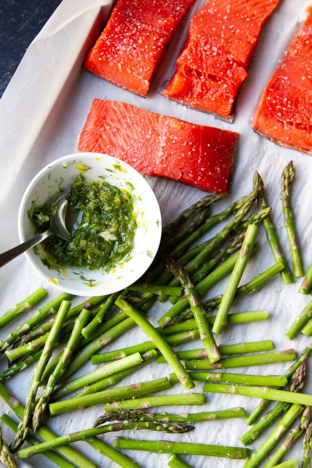 Parchment covered sheet pan with raw salmon fillets, uncooked asparagus, and a small bowl of herbs, garlic, & lemon zest mixture.