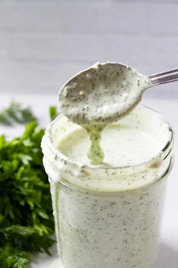 Finished green goddess dip in a glass jar with some of the dip running of a spoon back into the jar.