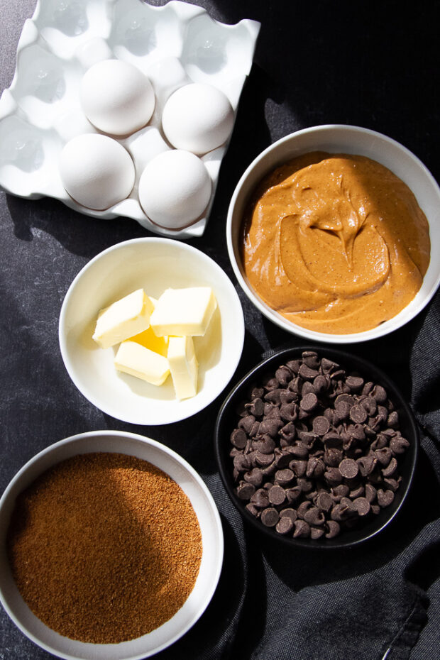 Ingredients for the chocolate peanut butter brownies in bowls -- eggs, peanut butter, butter, chocolate, and coconut sugar.