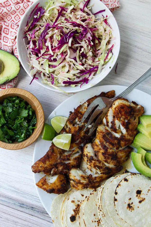 Cooked blackened fish on a plate with some sliced avocado and toasted corn tortillas. Next to a bowl of cabbage slaw and some fresh chopped cilantro.
