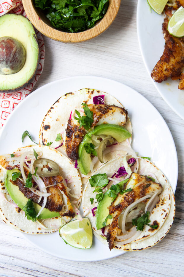 Three blackened fish tacos on a plate, dressed with avocado, cabbage slaw, fresh cilantro, and pickled onions.