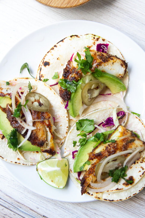 Three prepped blackened fish tacos on a plate. Dressed with avocado slices, cabbage slaw, and pickled onions.