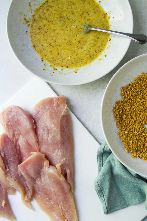 Thin, raw chicken breast pieces on a cutting board. Next to shallow dishes with wet & dry breading ingredients.