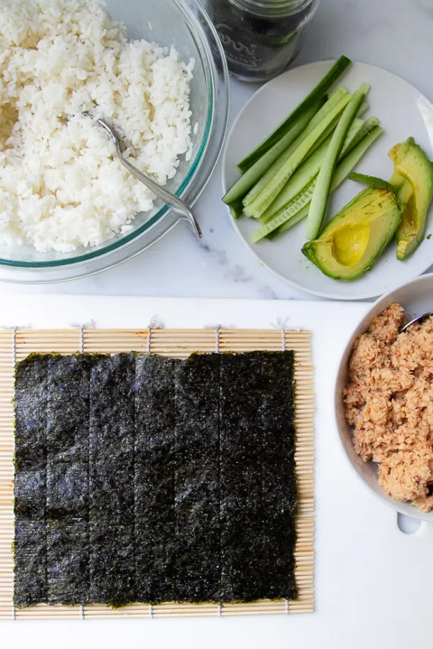 Workspace ready to roll up some sushi -- bamboo mat with a nori wrapper, bowls of spicy shrimp salad and sushi rice and a plate of thinly sliced cucumbers and avocado,.