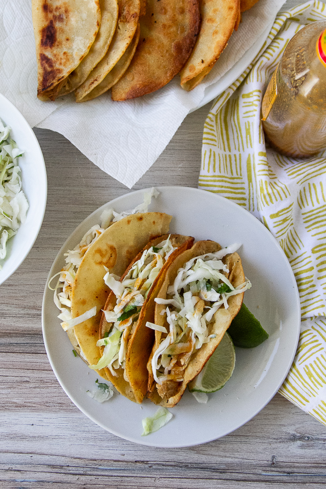 Crispy Chicken Tacos on a white plate with grated cabbage and lime wedges.