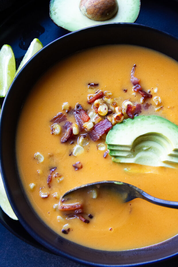 Butternut squash and sweet potato soup in a black bowl with corn, bacon, and avocado garnish.