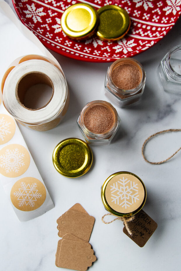 Tiny jars with cinnamon sugar being packaged up with gold lids and snowflake stickers on the lids. Also shown: twine, tags, and stickers.