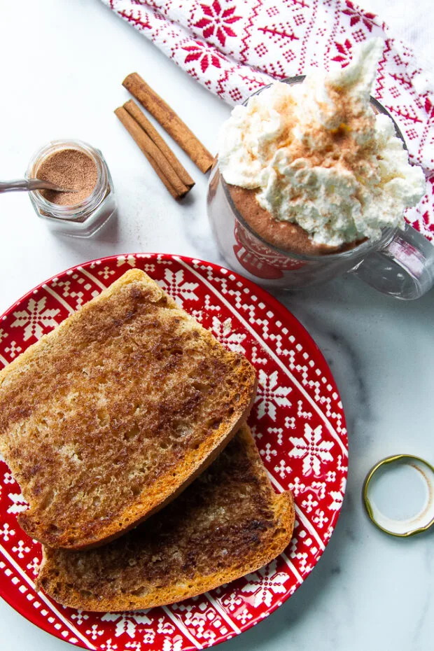 A plate with two slices of cinnamon toast and a mug of hot chocolate with whipped cream topped with some cinnamon sugar.