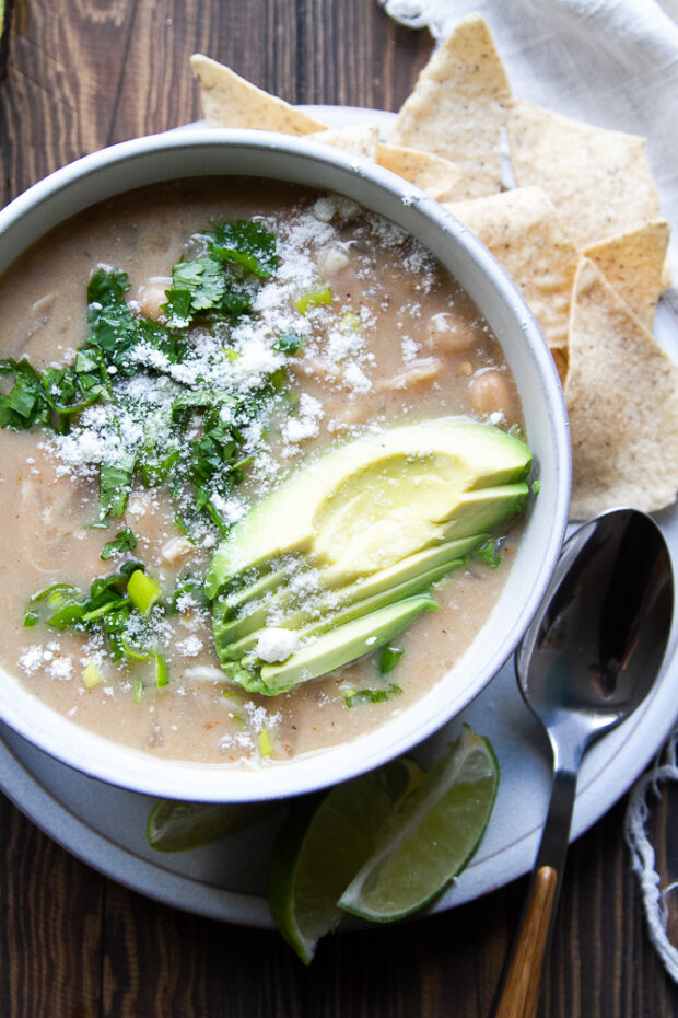White chicken chili in a gray bowl topped with sliced avocado, fresh cilantro, green onions, and cotija cheese. Tortilla chips and lime wedges on the side.