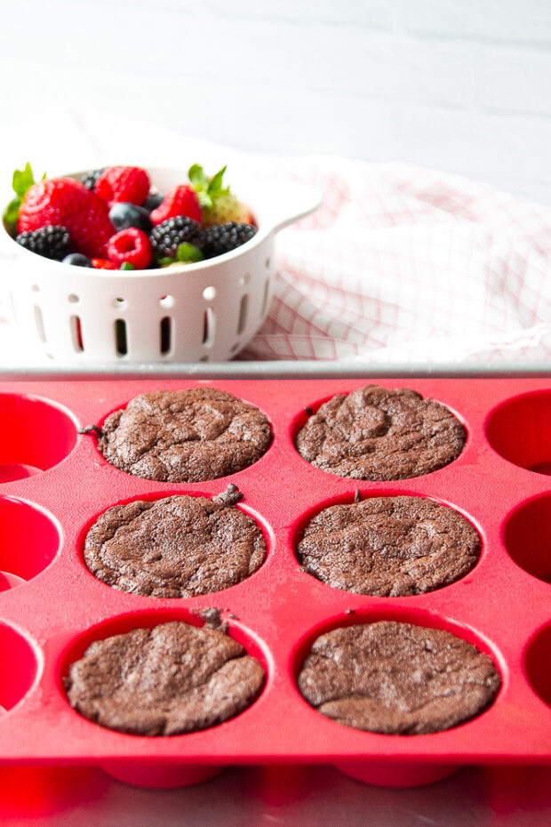 Freshly baked flourless chocolate cakes still in the muffin pan. A bowl of berries sits next to it.
