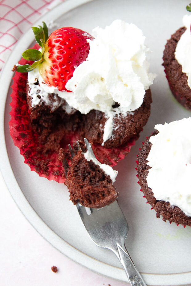 A brownie version of the flourless chocolate cakes with whipped cream and strawberries. A fork has taken a chunk out of one of the cakes.