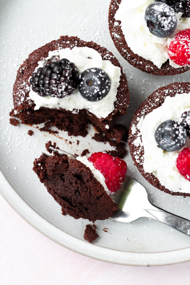 Three individual flourless chocolate cakes on a plate dressed with some whipped cream, berries, and a dusting of powdered sugar. A fork has take a bit-sized piece out of one.