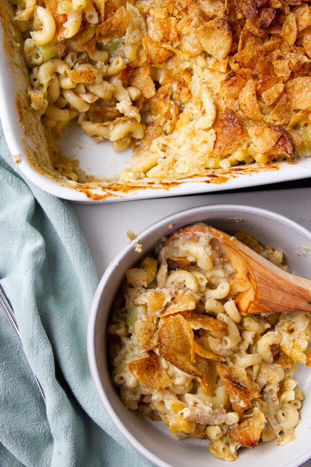 Casserole dish with baked tuna noodle casserole. One serving has been scooped out into a bowl with a wooden serving spoon.