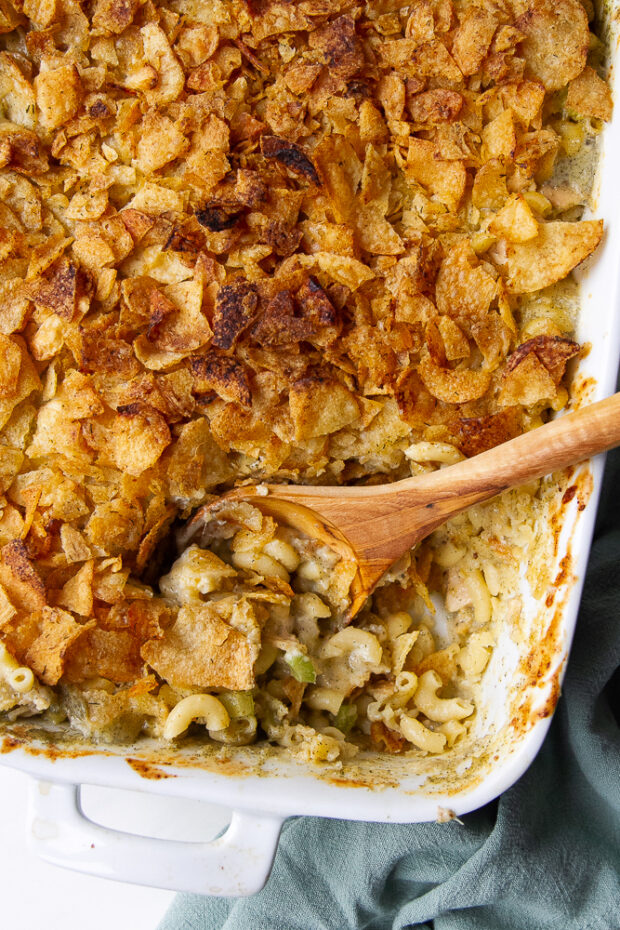 Tuna noodle casserole in a white baking dish with a wooden serving spoon stuck into the corner. A serving of pasta has been removed from the pan.