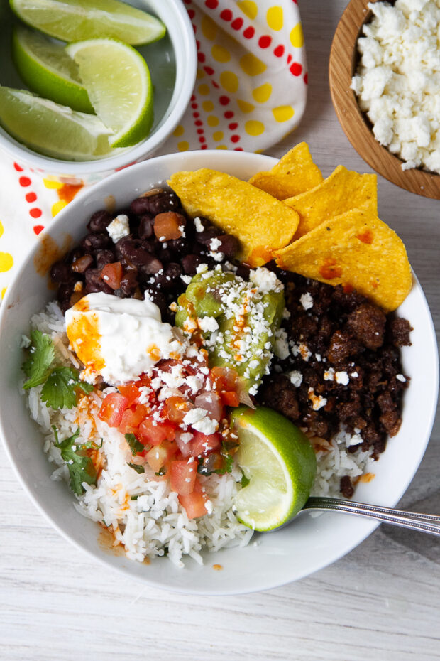A burrito bowl assembled in a white bowl with smaller bowls of lime wedges and cotija cheese off to the side.
