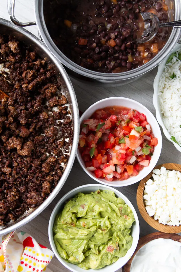 Components for burrito bowls displayed in their individual containers: ground taco meat, black beans, pico de Gallo, rice, cotija cheese, sour cream, and guacamole.