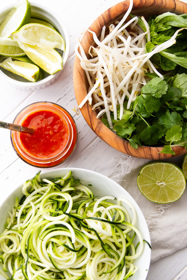 Garnishes and add-ins for the soup set out: lime wedges, sambal oelek, zoodles, mung bean sprouts, fresh cilantro, fresh basil, and limes.