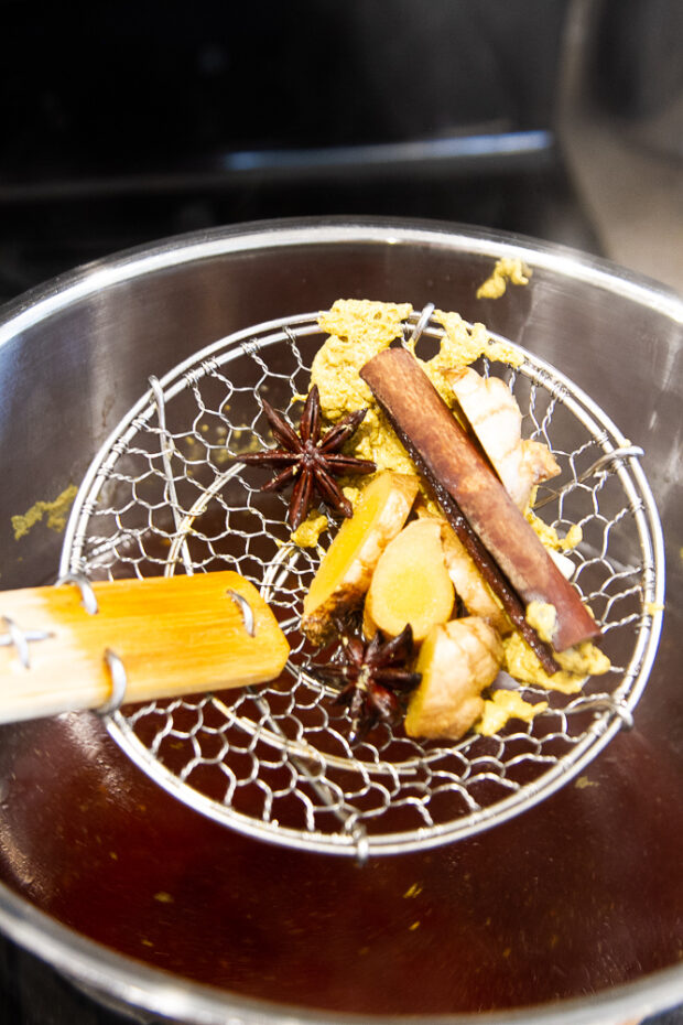 A strainer puling the star anise pods, cinnamon sticks, and ginger out of the broth.