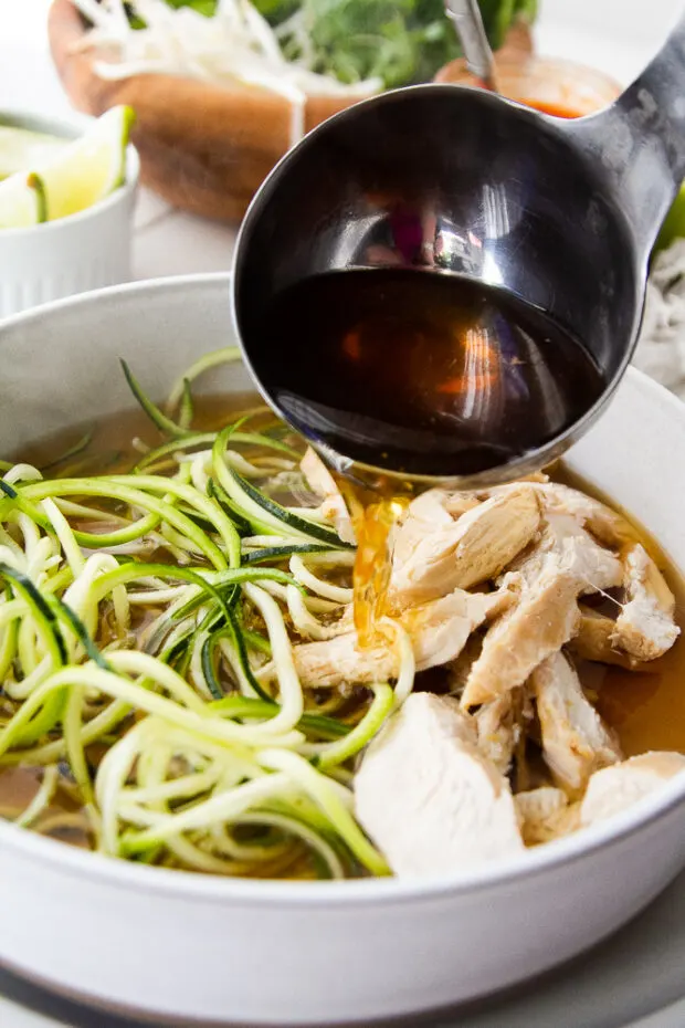 A ladle pouring hot broth into a bowl with zoodles and cooked chicken.