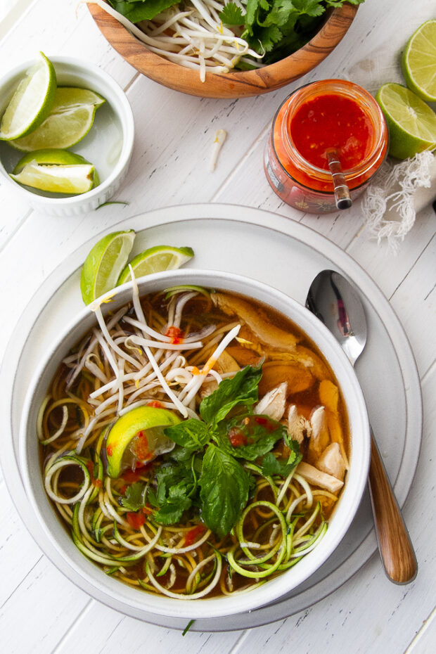 Bowl of Chicken Pho soup with garnishes on top and the garnishes set around the bowl.