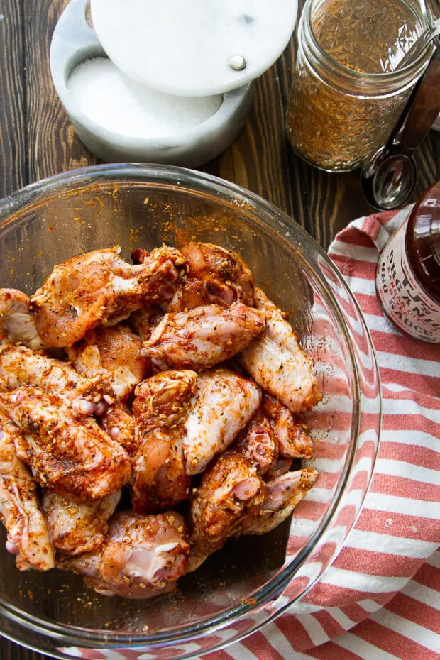 Raw chicken wings in a clear bowl that have been tossed in oil and seasoning.