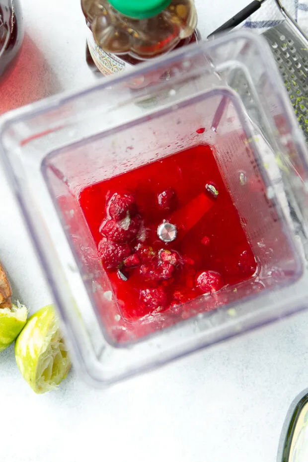 Overhead view of the dressing ingredients in a blender ready to blend: thawed frozen raspberries, lime juice, lime zest, honey, red wine vinegar, and avocado oil.