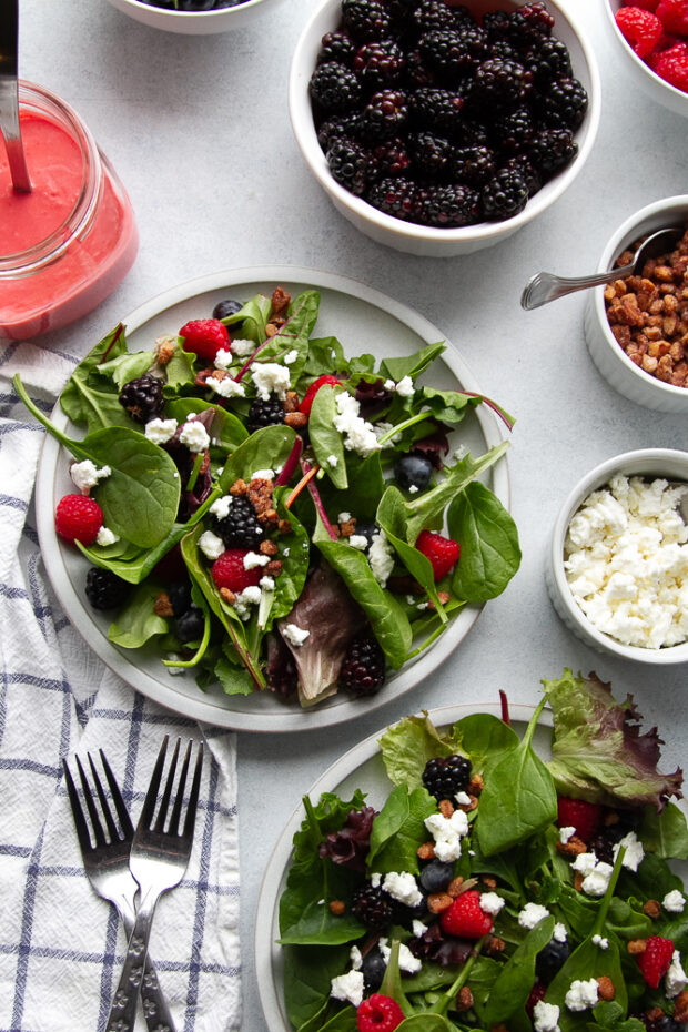 An assembled salad without dressing on a white plate. The toppings are arranged around in smaller bowls and the dressing is off to the side.