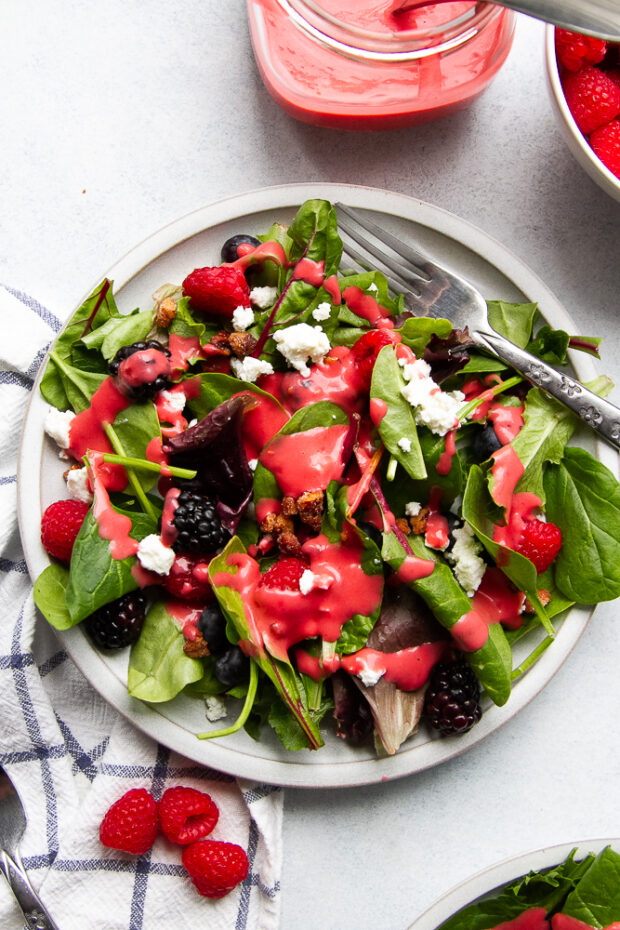A spring mix salad on a white plate topped with raspberry vinaigrette, fresh berries, goat cheese, and candied nuts. 