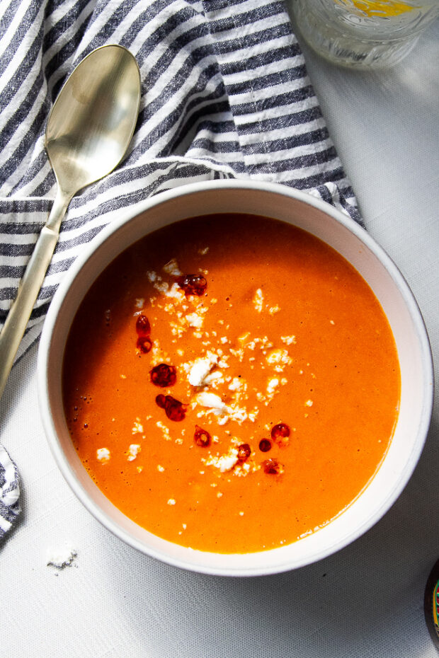 A bowl of red pepper tomato soup in a white bowl with goat cheese and chili crunch as garnish.