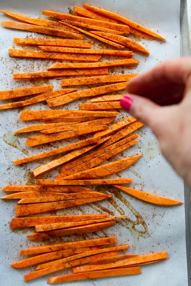 Uncooked sweet potato fries on a parchment lined baking sheet. Drizzled with oil and sprinkled with seasoning.