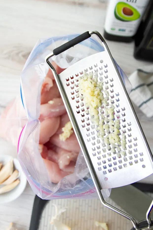 Raw chicken in a ziptop bag with a Microplane grater adding grated garlic to the bag.