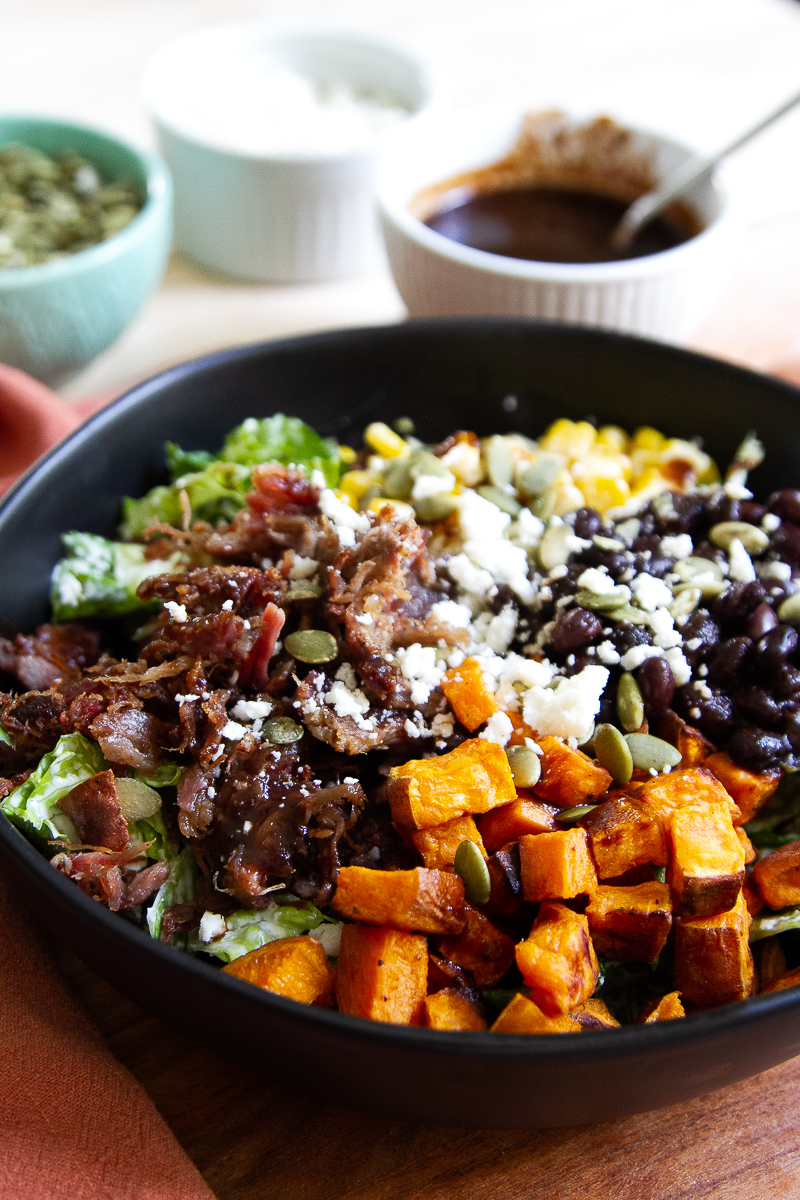 Black bowl with BBQ pork salad. Romaine lettuce tossed in jalapeno ranch, roasted fresh corn, roasted sweet potatoes, crispy pulled pork, black beans, pepitas, cotija cheese, and bbq sauce drizzle.